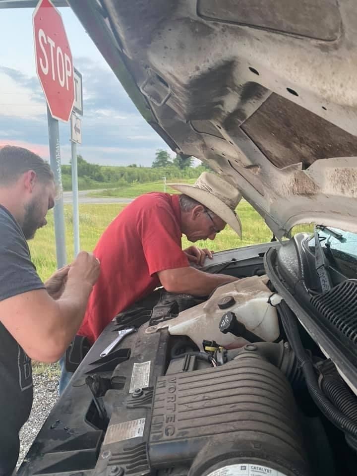 Two men working on the engine of a van, one wearing a cowboy hat, next to a stop sign.