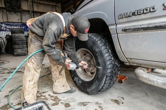 Mechanic uses an impact wrench on a truck's wheel. He wears dirty work clothes in a garage setting.