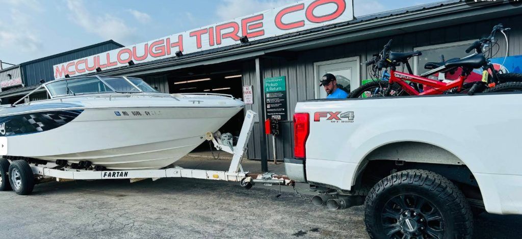 A boat on a trailer being pulled by a white truck in front of a tire shop.