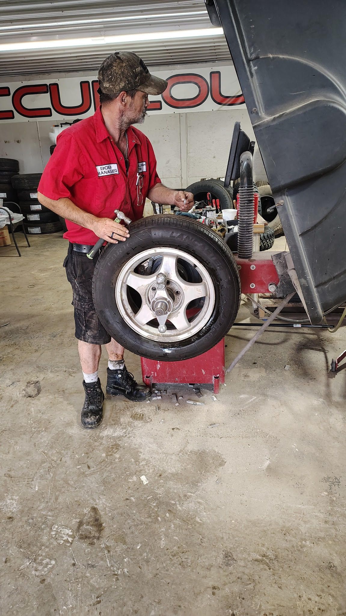 Mechanic holding a car tire near a wheel balancer in a garage setting. He wears a red shirt, camo hat.