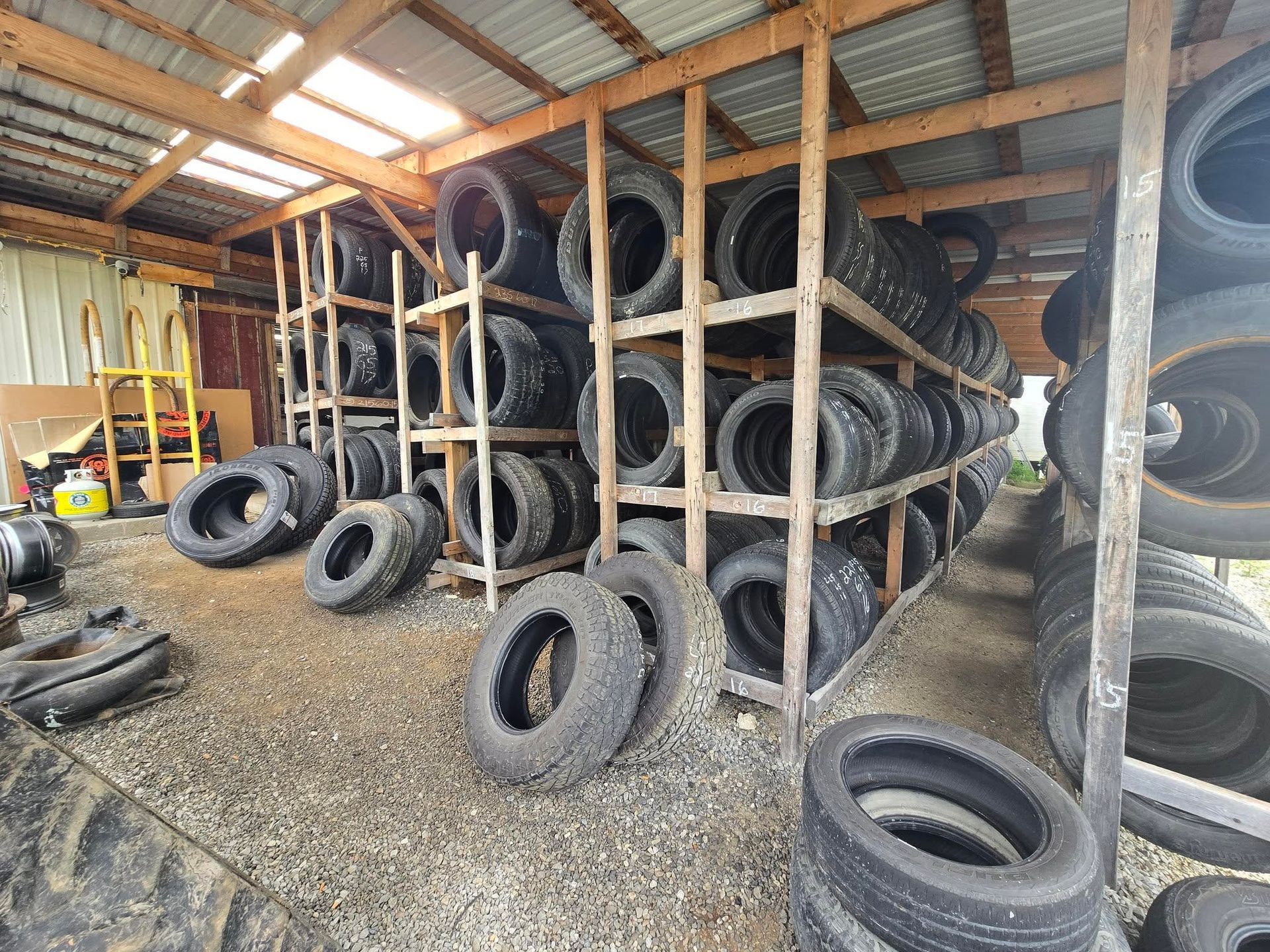 Tires stored on wooden shelves inside a shed. Some tires are on the floor. Gray gravel ground.