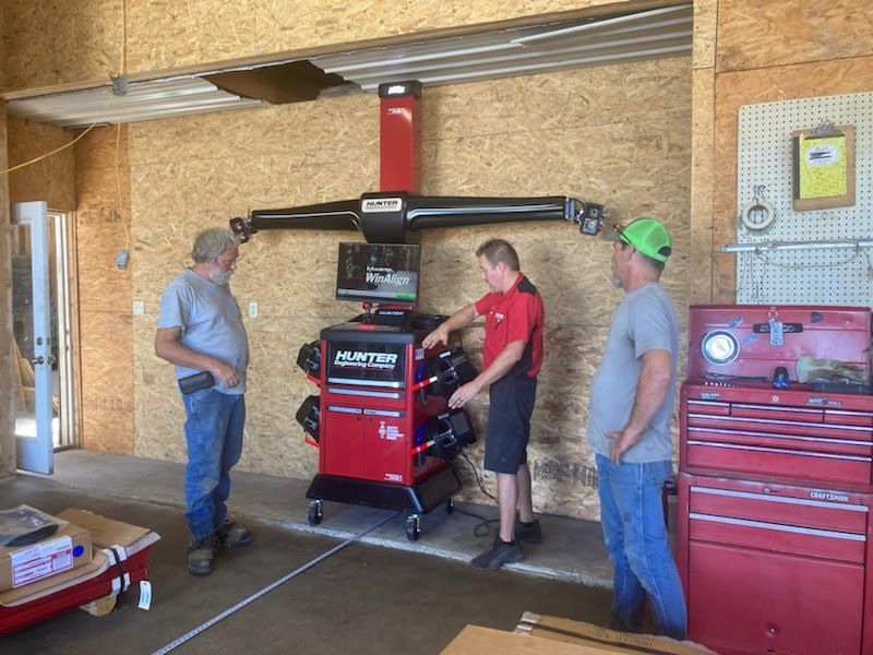 Three men in a garage setting with a wheel alignment machine; one man points.