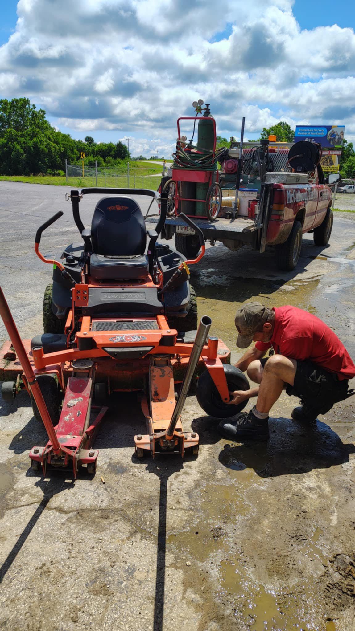 Man changing tire of an orange zero-turn mower, jack stands. Red truck in background. Sunny, outdoor setting.