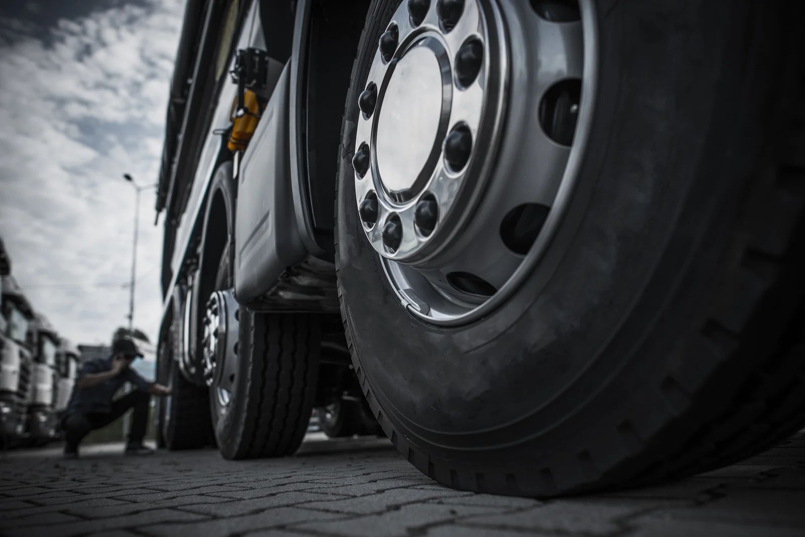 Close-up of a large truck wheel on a cobblestone street, with a person in the background and cloudy sky.