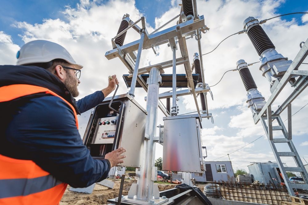 Engineer in a hard hat and orange vest working on electrical equipment outdoors.