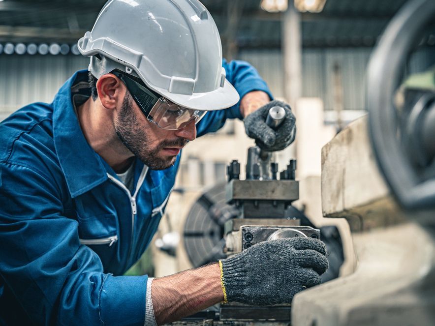 Mechanic in blue coveralls, safety glasses, and hard hat, working on machinery in a factory.