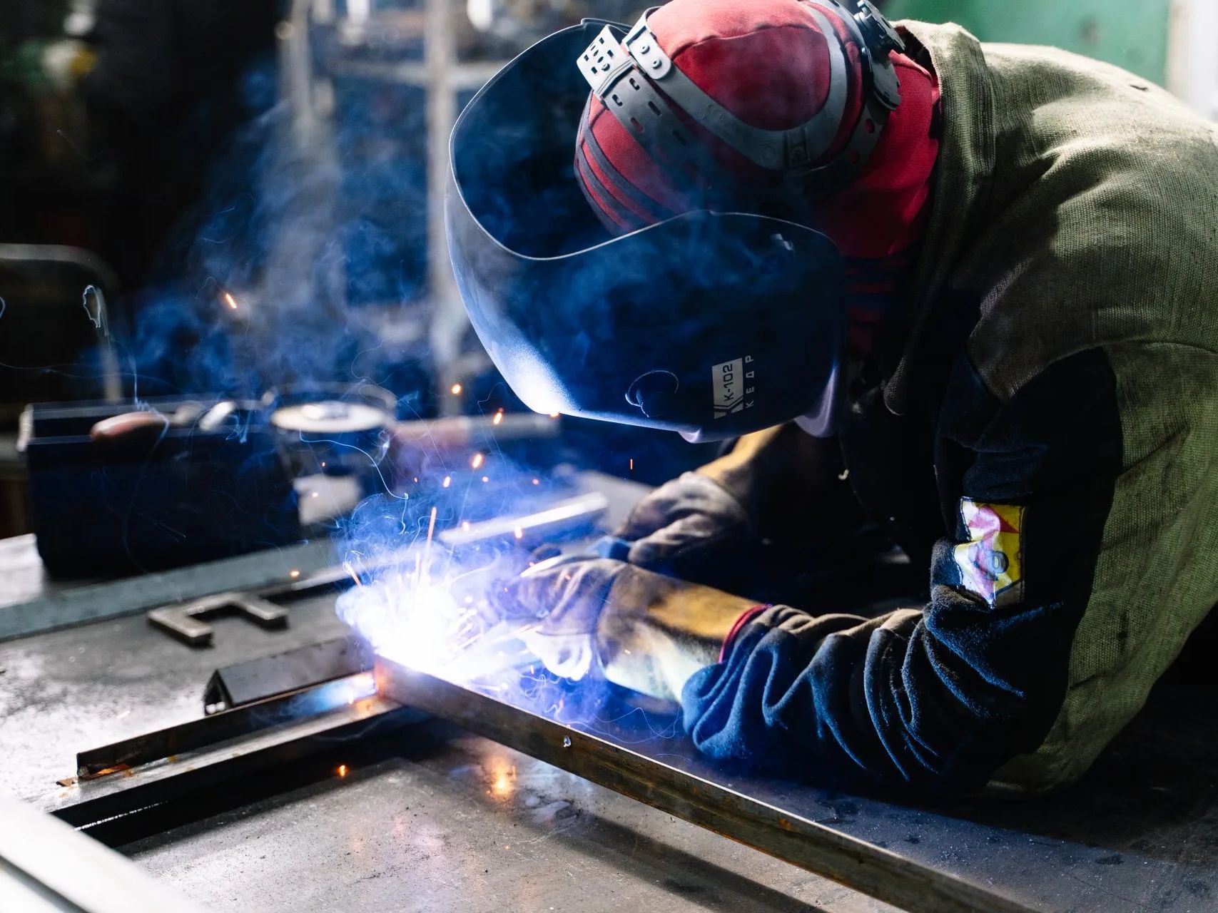 Welder wearing a protective helmet, welding metal with bright sparks in a workshop.