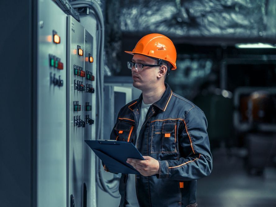 Man in orange hard hat and work clothes examining electrical panel, holding clipboard.