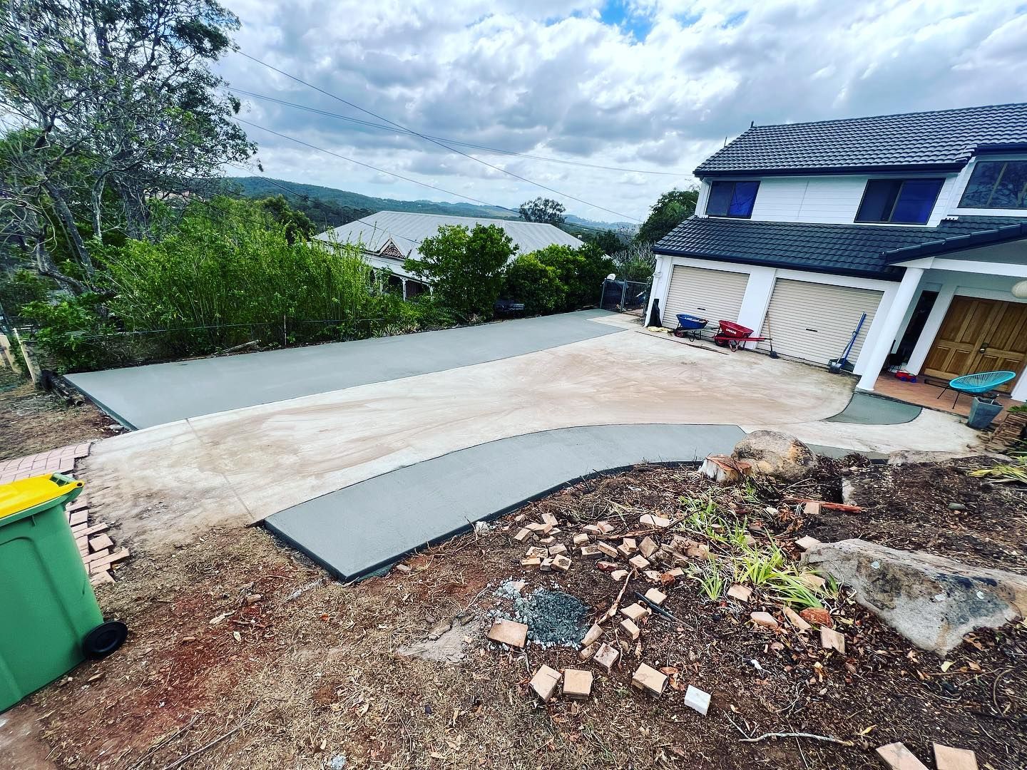 Concrete driveway beside a blue house under construction, with gravel, plants, and a hillside view.