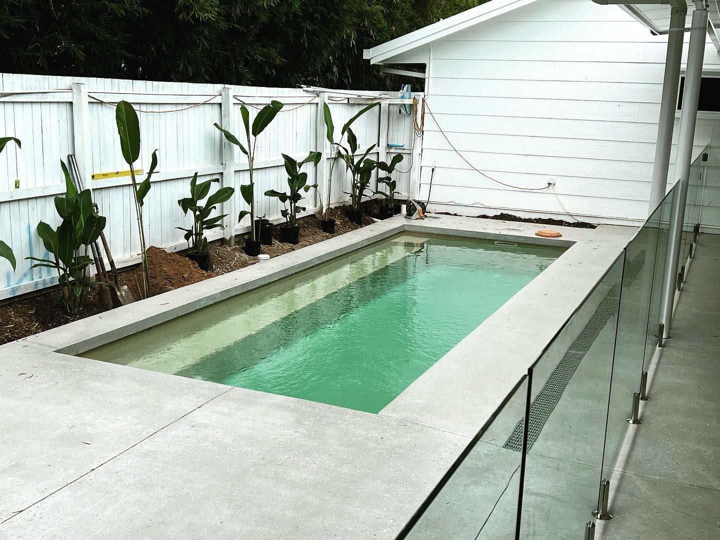Narrow backyard lap pool beside a white fence and garage, with tropical plants along the edge.