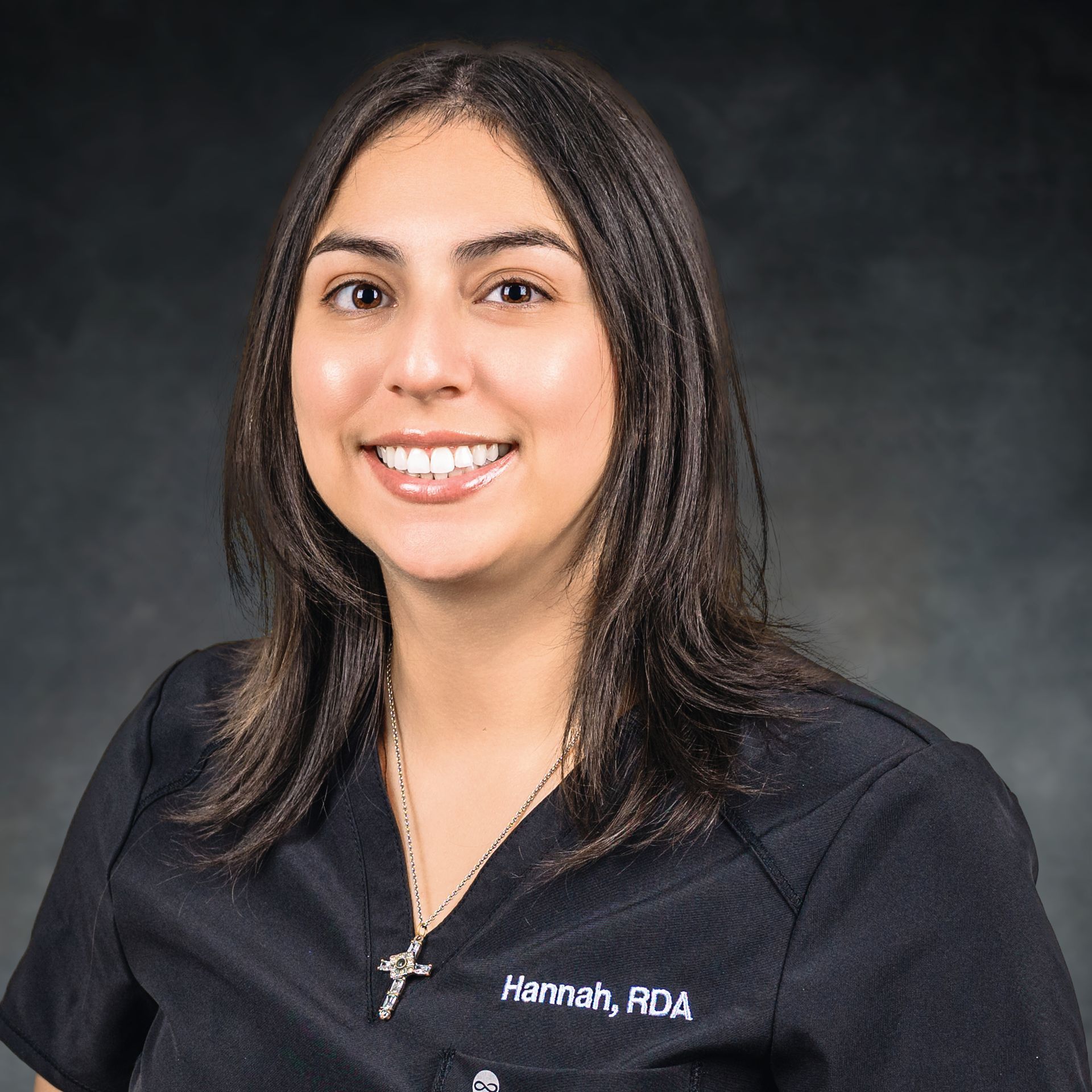 Woman smiling, wearing black scrubs, a necklace, and a nametag that reads