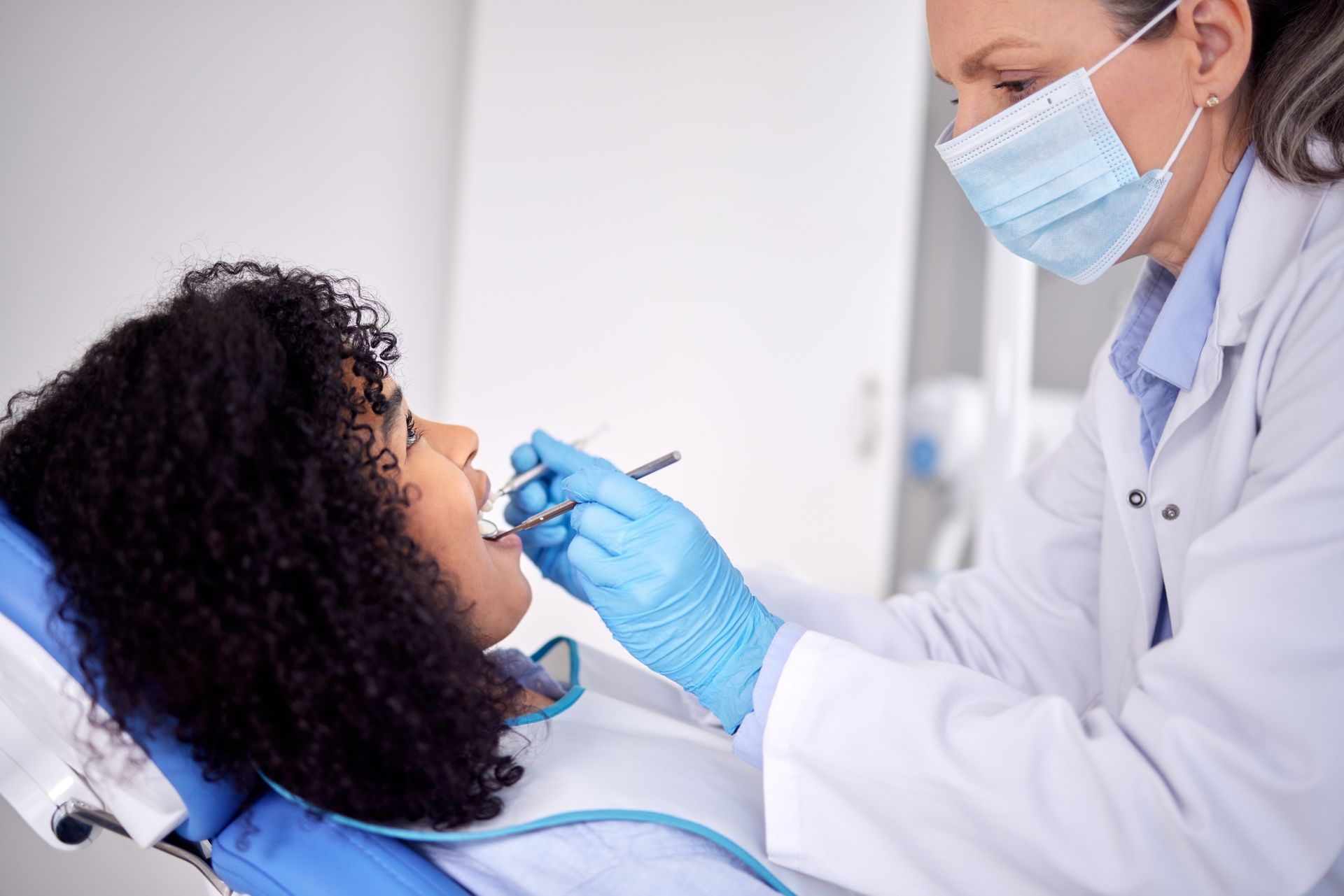 Dentist in mask and gloves examining a patient’s teeth in a dental chair.