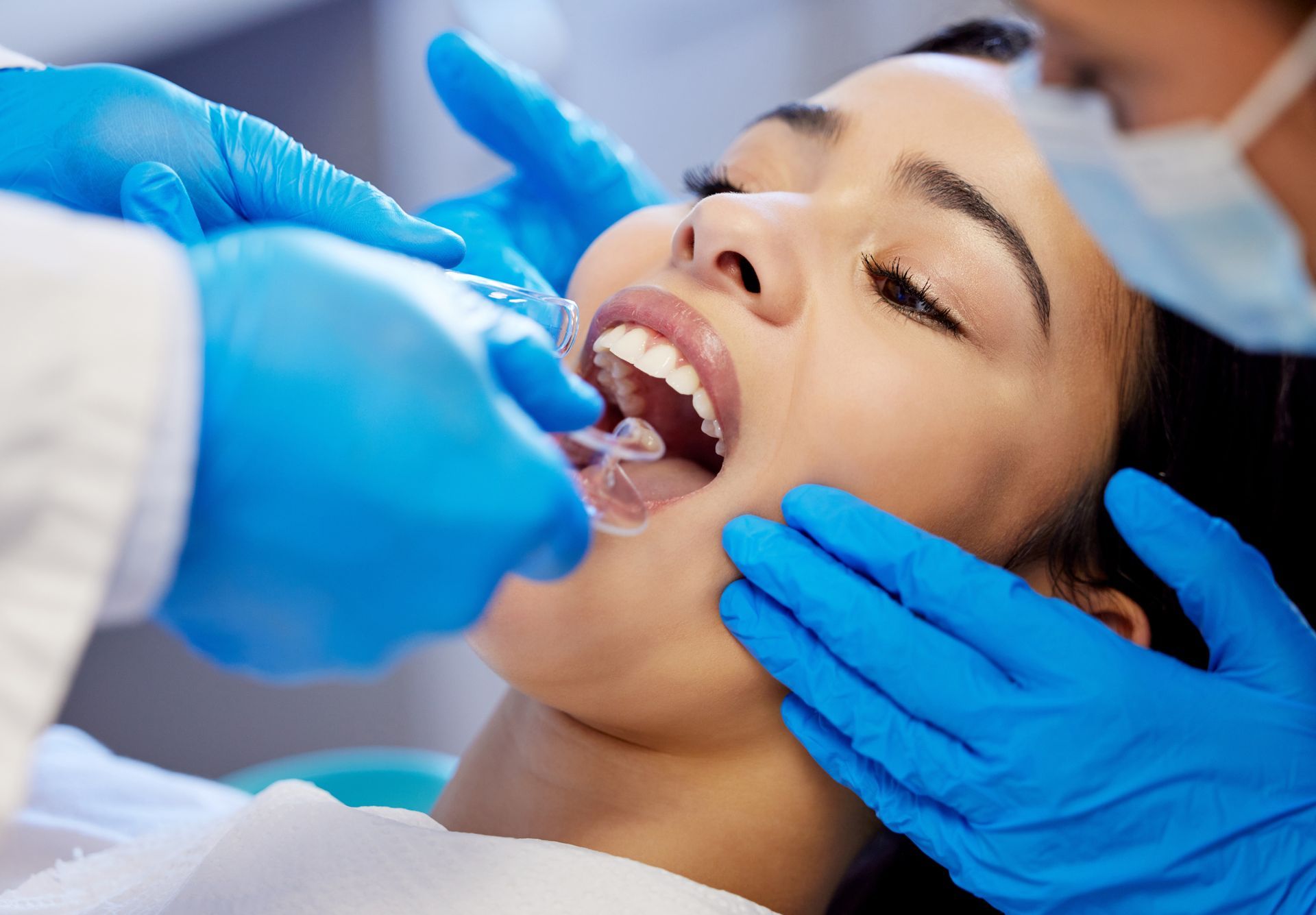 Woman at dentist, mouth open, receiving dental care by two people wearing gloves and masks.