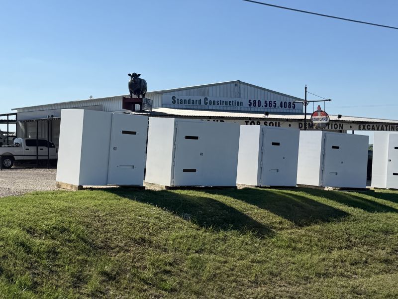 White above ground storm shelter units in a grassy field beside a building under a clear blue sky