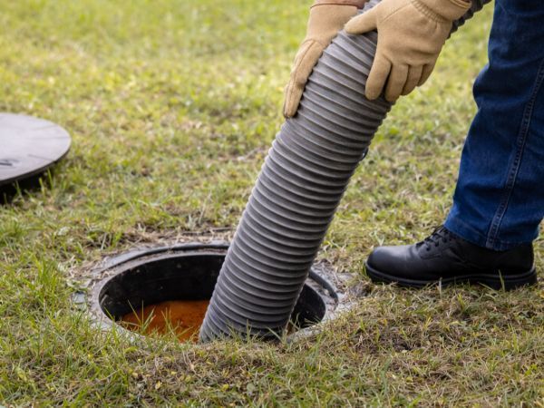 Person inserting a gray pump hose into a round septic tank on a grassy lawn