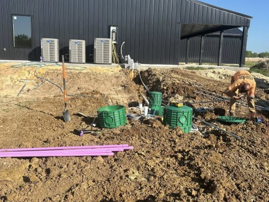 Construction site with muddy trench, green utility boxes, purple pipes, and a worker near a black building