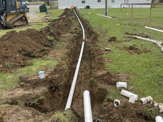 A long white pipe lies in a trench across a grassy yard, with dirt piles and pipe fittings scattered nearby.