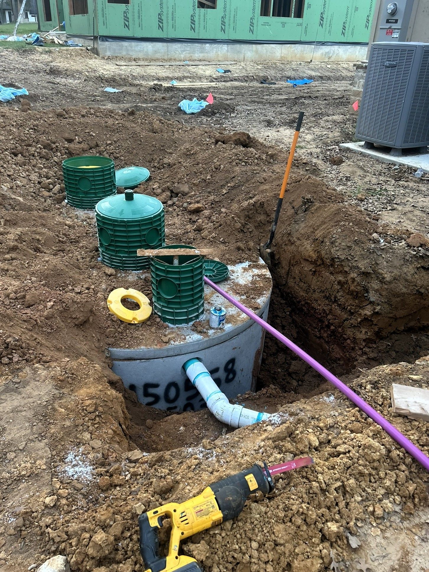 Construction trench with exposed pipes, green utility covers, and a yellow drill on muddy ground