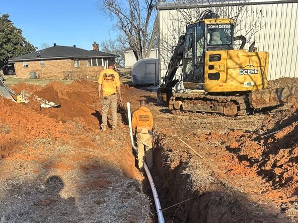 Construction crew working in a dirt trench beside a yellow excavator near houses