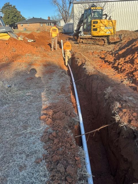 Construction trench with a white pipe being laid, workers in orange vests, and an excavator on a dirt site
