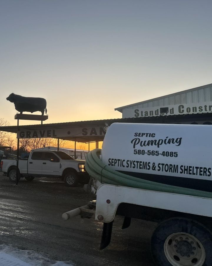 A septic pumping truck parked in front of a construction business with a metal cow silhouette on the roof at sunset.