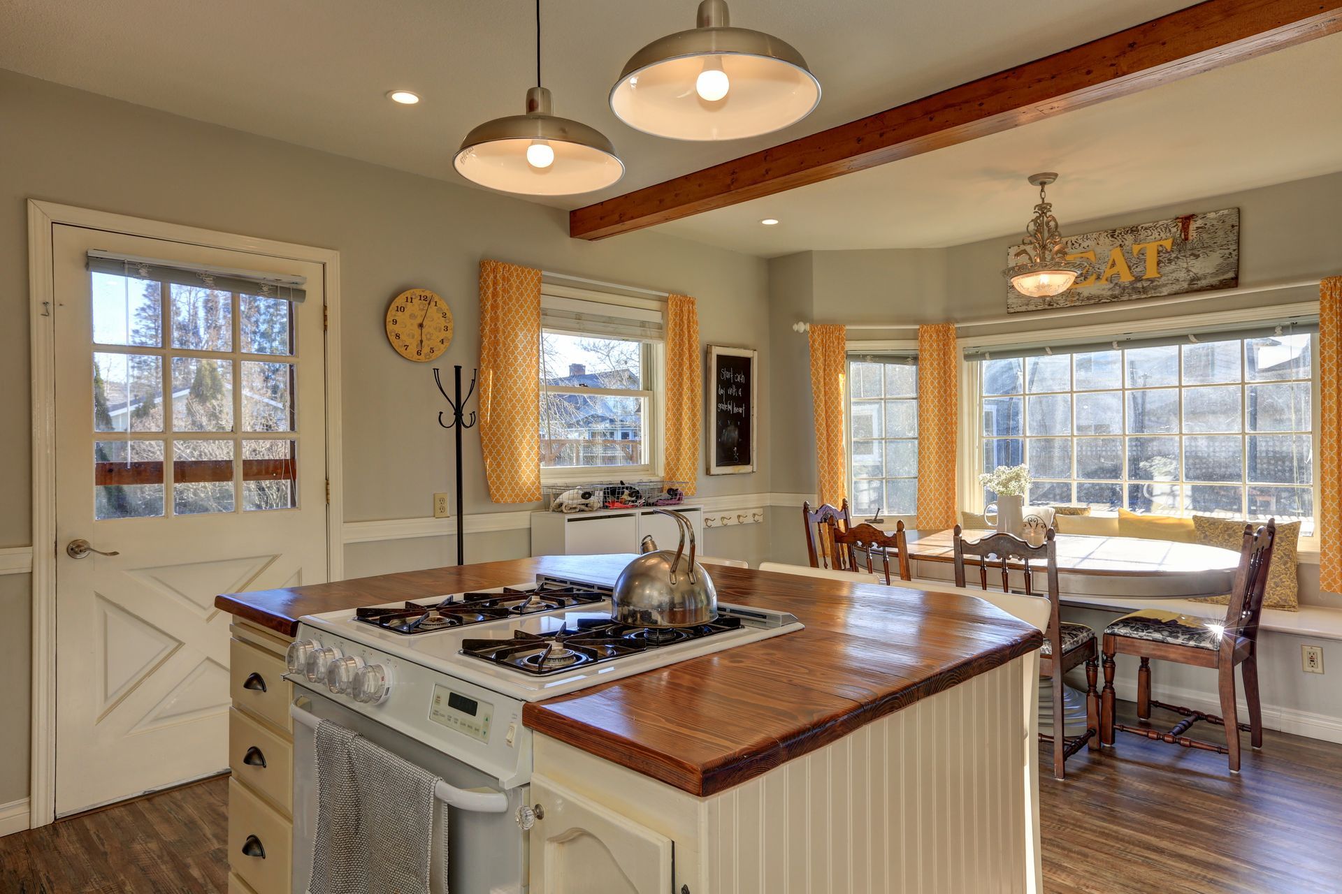 Cozy kitchen with a stove island, wooden beams, and a bay window seating area with yellow curtains.