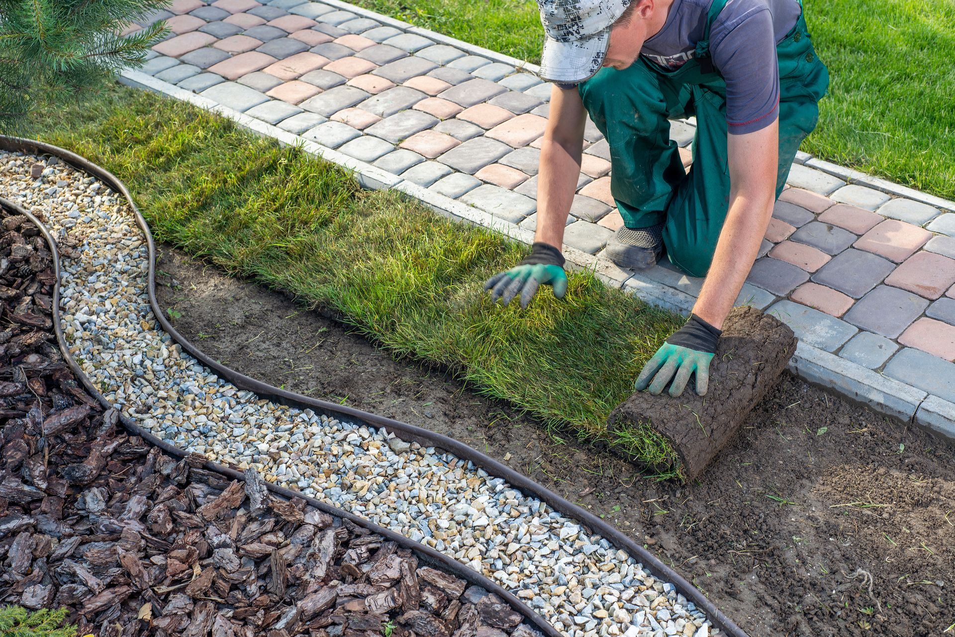 Man laying sod near a walkway. He kneels, wearing gloves, placing a roll of grass next to a gravel border.