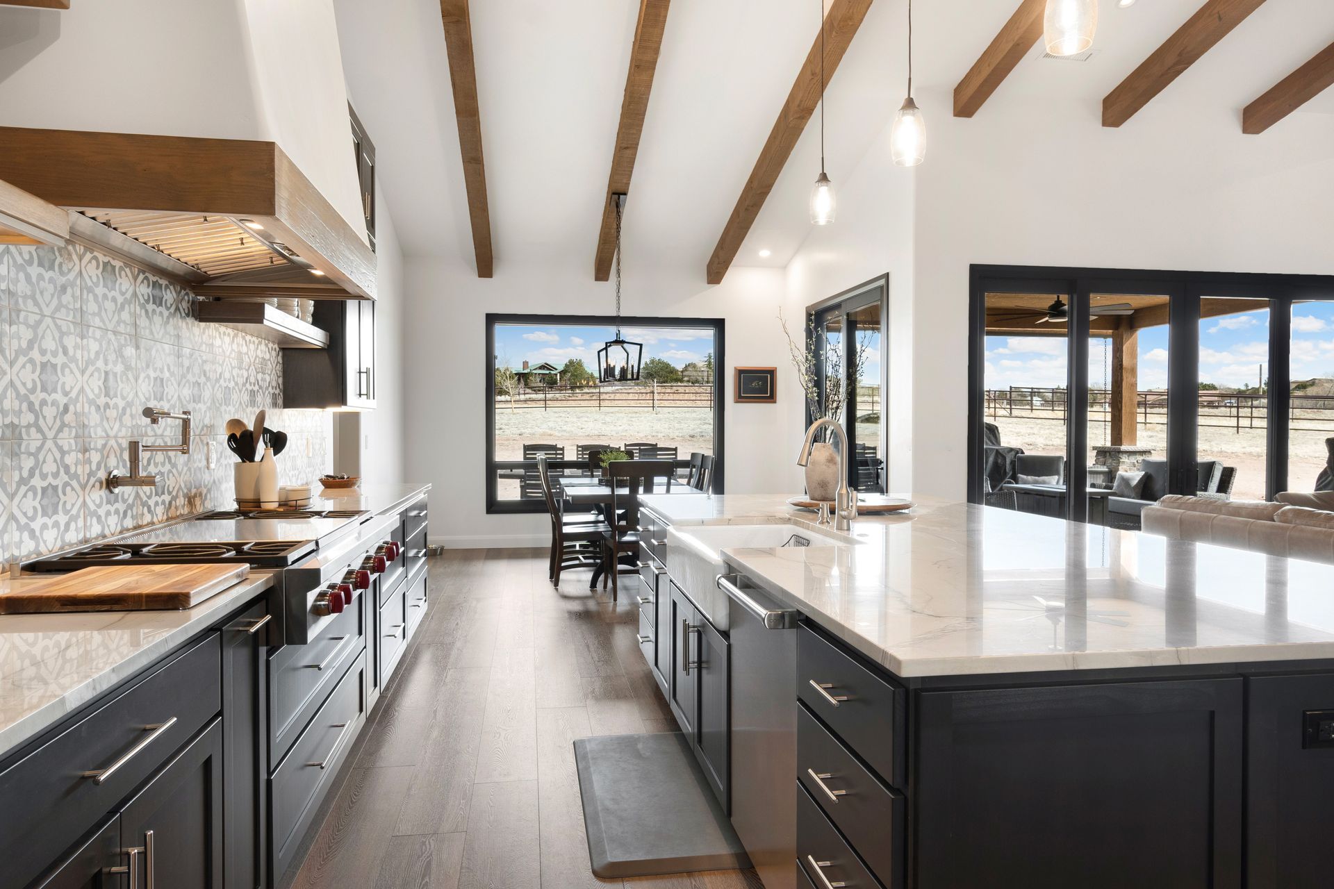 Modern kitchen with dark cabinets, white countertops, and wooden ceiling beams, open to a dining area and outdoor patio.