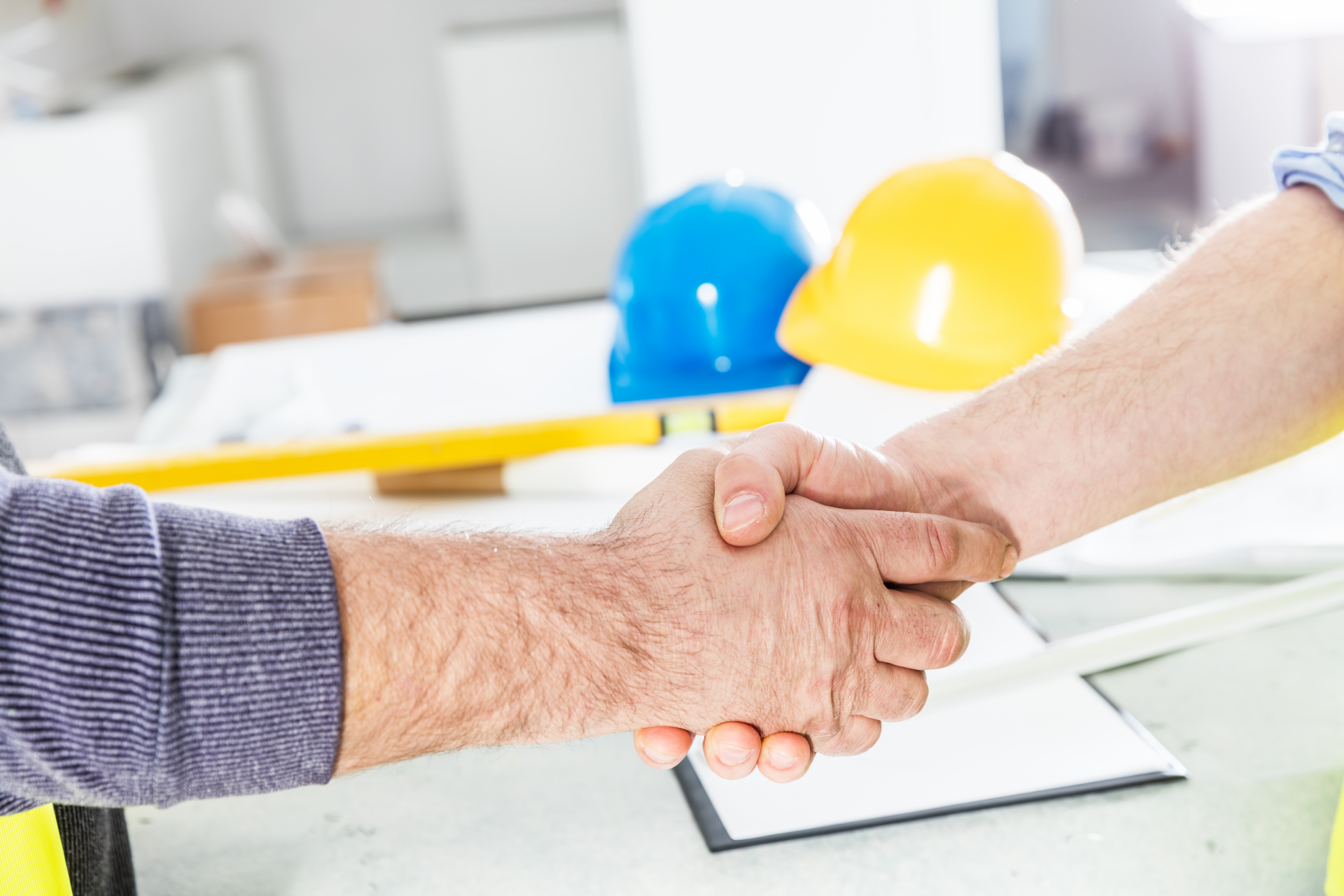 Two Construction Professionals shaking hands in front of a desk with hard hats and constructin plans on the desk
