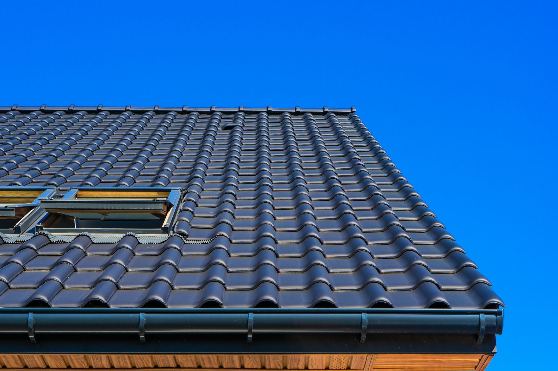 A low-angle view of a tiled roof with a skylight against a clear, solid blue sky.