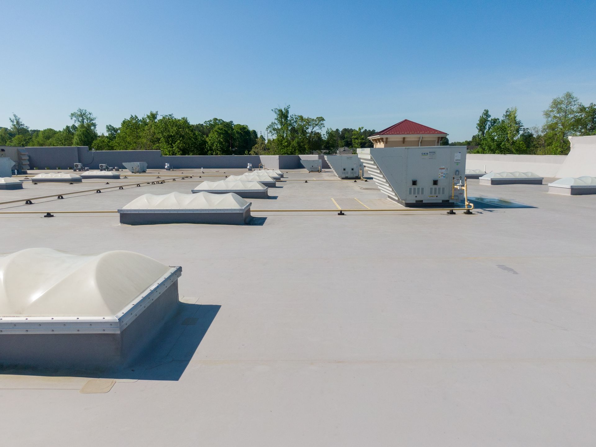 Flat rooftop with white vents and skylights under a clear blue sky, with trees in the distance.