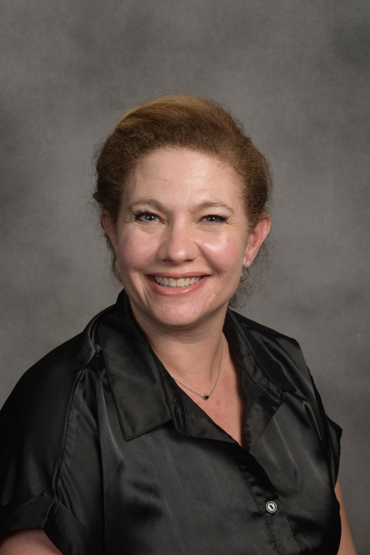 Woman with auburn hair smiles, wearing a black satin shirt. Against a blurred gray background.