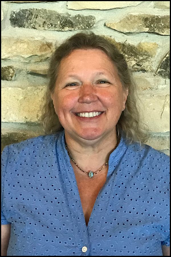 Woman smiling, wearing a blue dotted shirt, with a stone wall background.