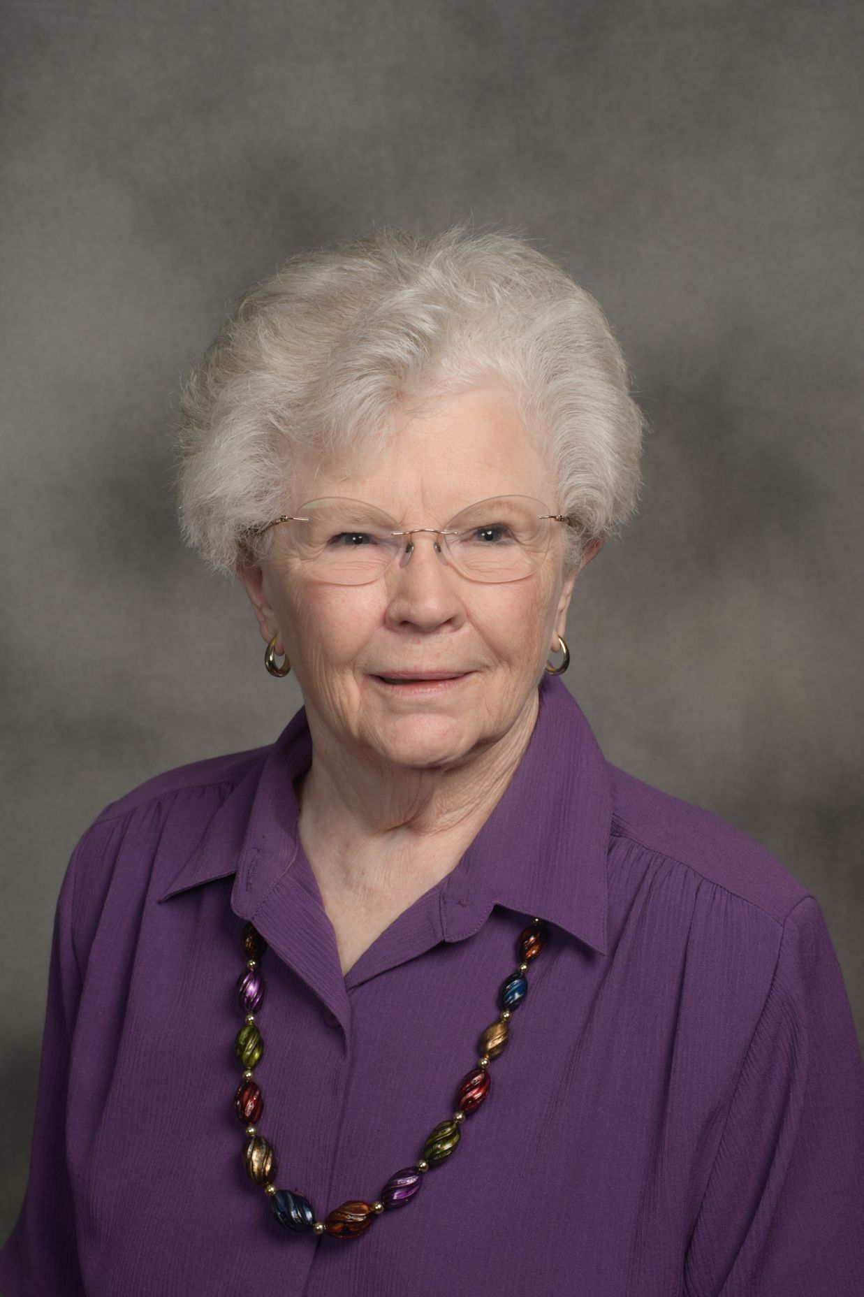 Woman with white hair and glasses wearing a purple shirt and beaded necklace, smiling.