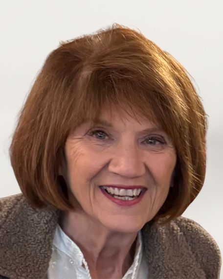 Woman with short auburn hair smiles widely, wearing a brown jacket over a white top.