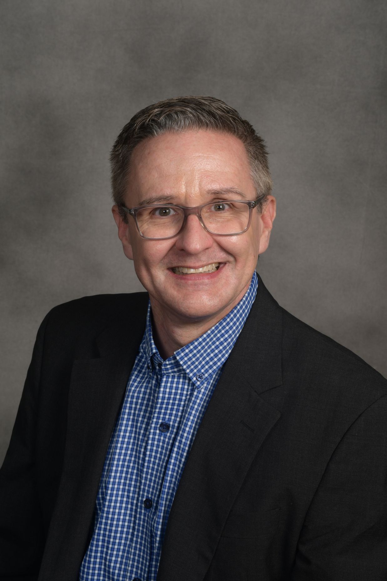Man wearing glasses, blazer, and checkered shirt, smiling against a gray background.