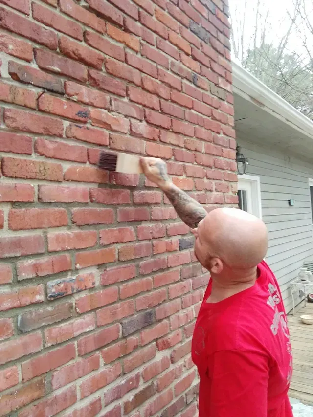 Man painting brick wall with a brush. Red shirt, shaved head, outdoor setting.