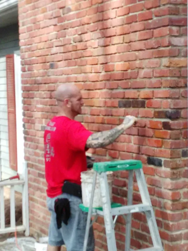 Person in red shirt on ladder, applying substance to brick wall.