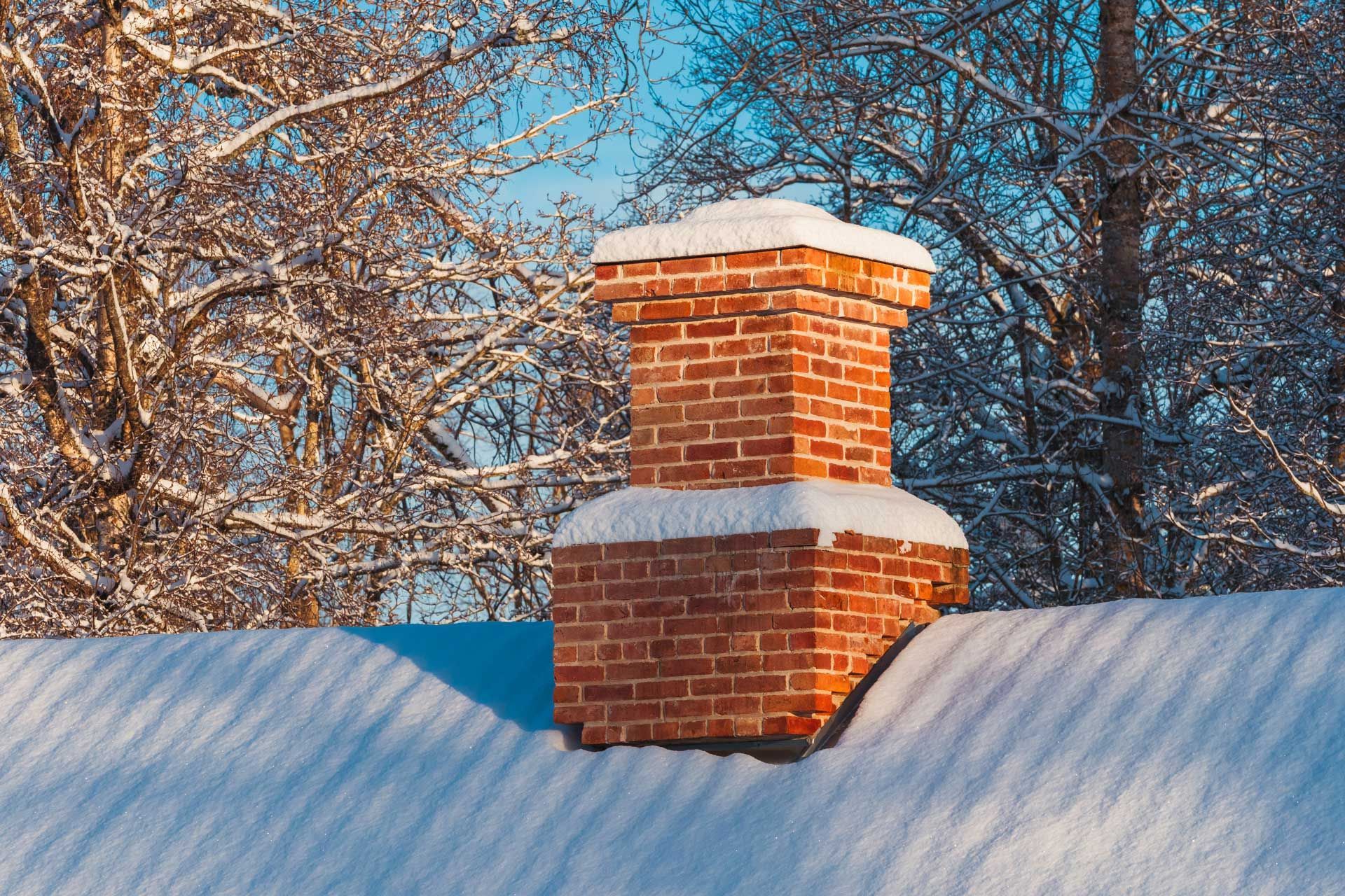 Brick chimney on a snow-covered roof, surrounded by snow-dusted trees under a blue sky.