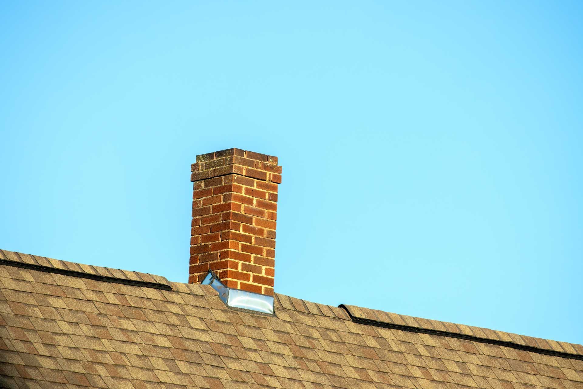 Brick chimney on a brown shingled roof against a clear blue sky.
