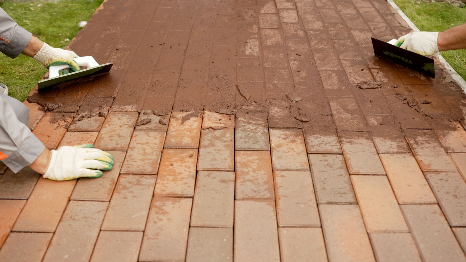 Two workers in gloves applying brown grout to brick paving with trowels.