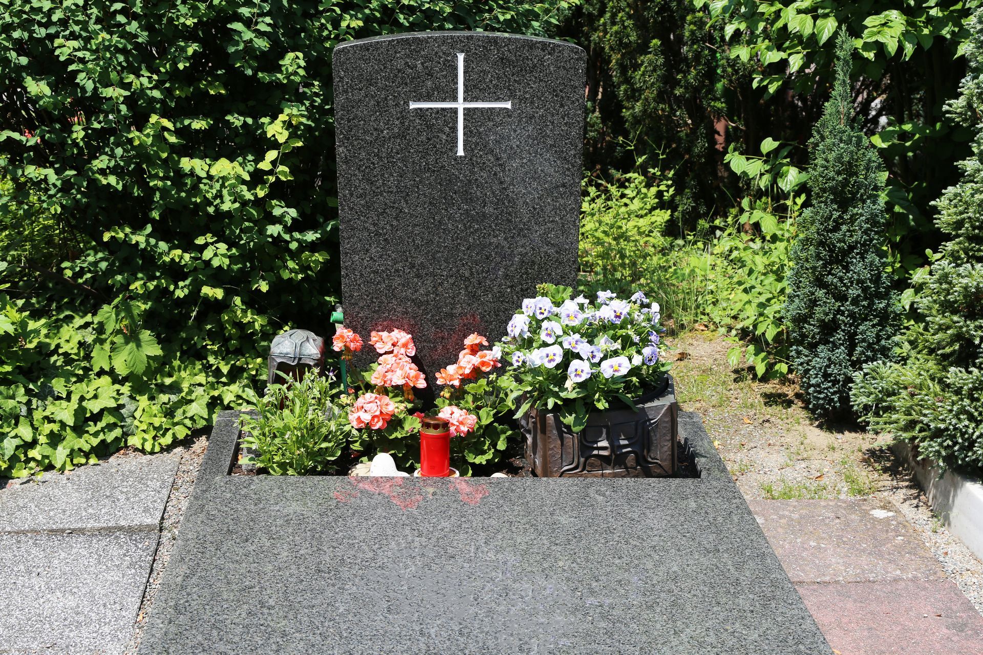 Grave with dark headstone and cross, flowers, and a red candle in a garden setting.