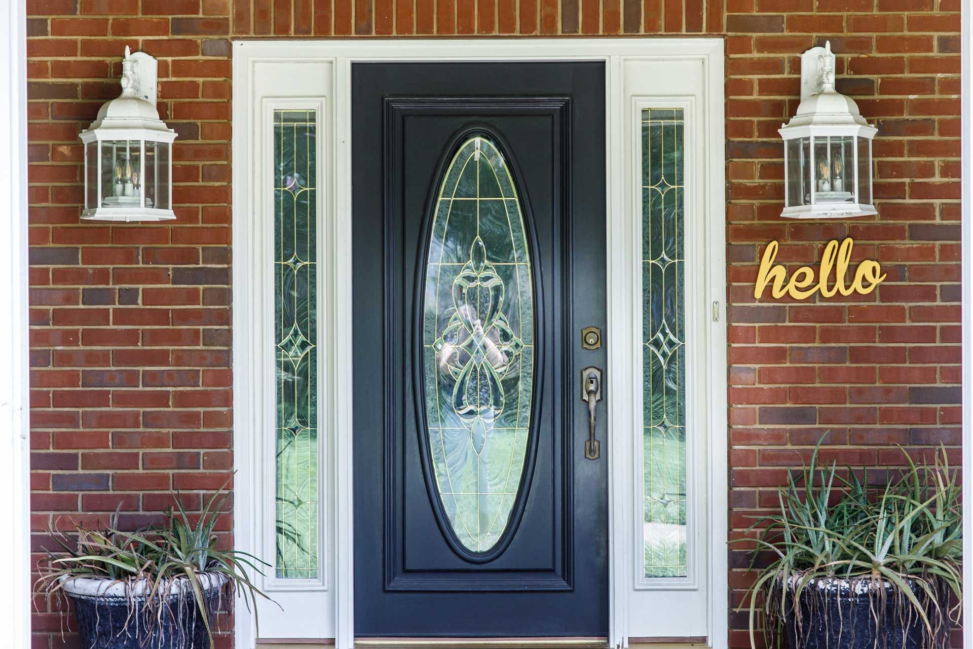 Black front door with oval glass panel, flanked by sidelights and lanterns, set in a brick facade, 