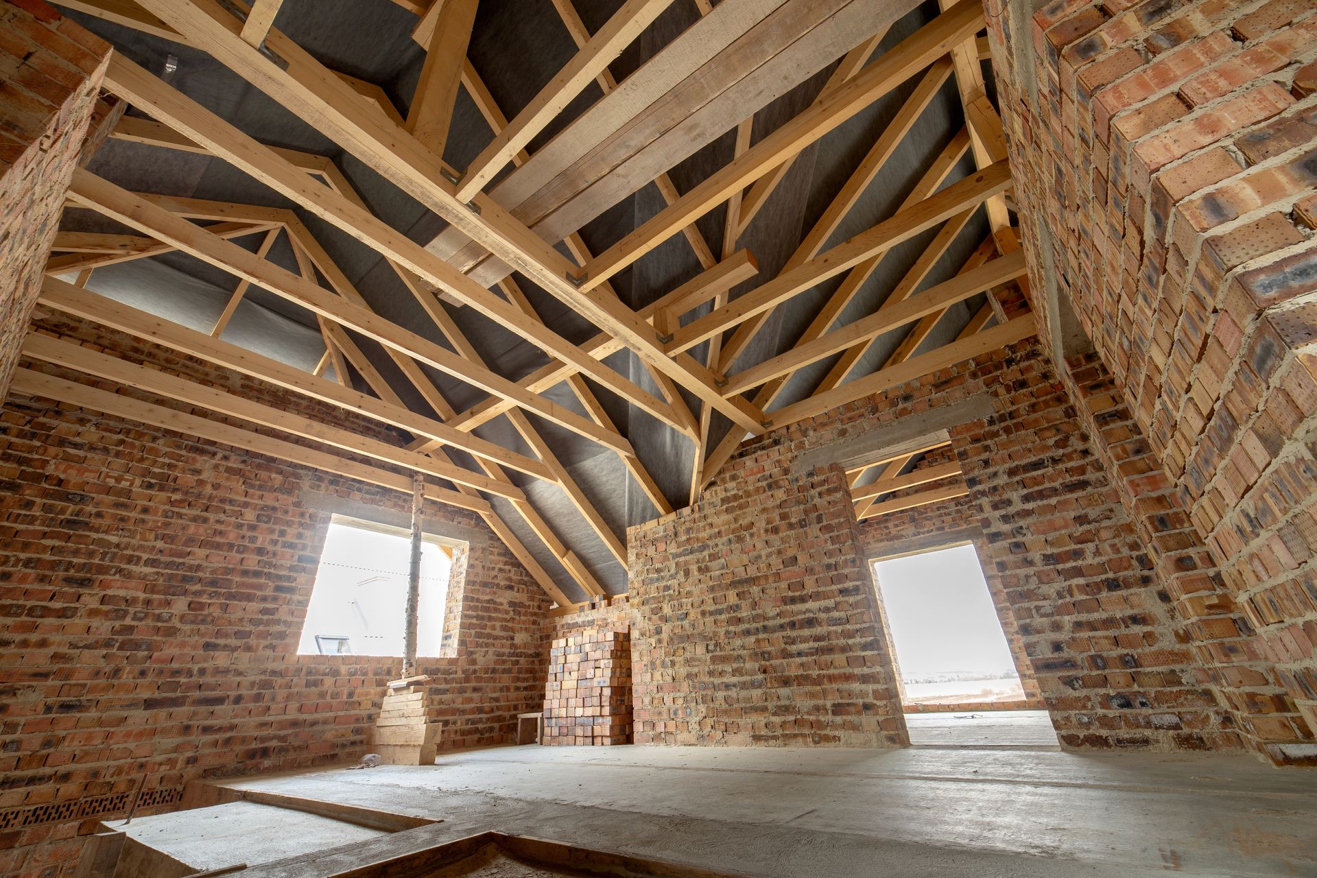 Interior view of a building under construction, showing exposed wooden roof beams, brick walls, and concrete floor. Interior view of a building under construction, showing exposed wooden roof beams, brick walls, and concrete floor.