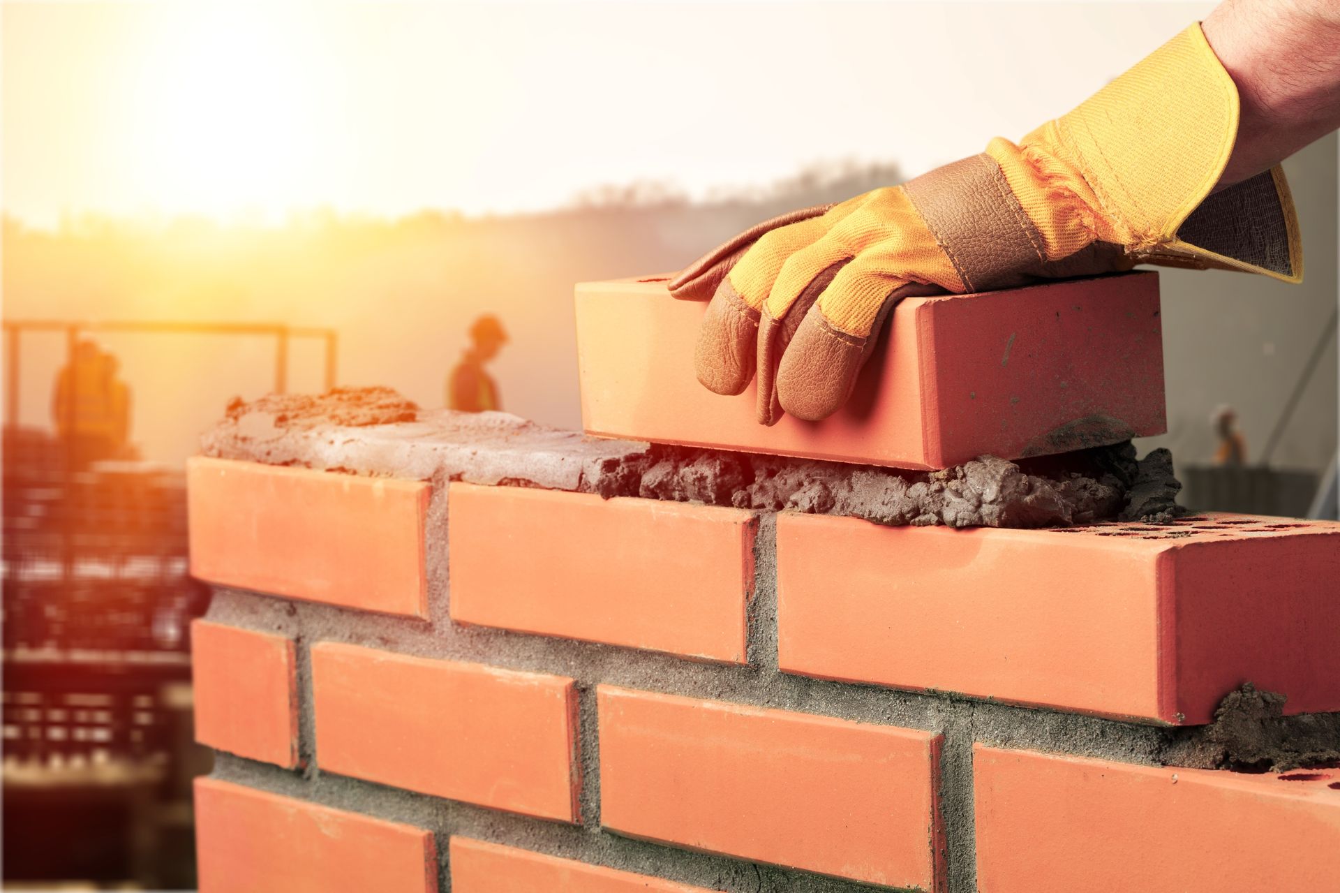 Bricklayer placing a brick on a partially built wall, wearing work gloves, outdoors with sunlight.