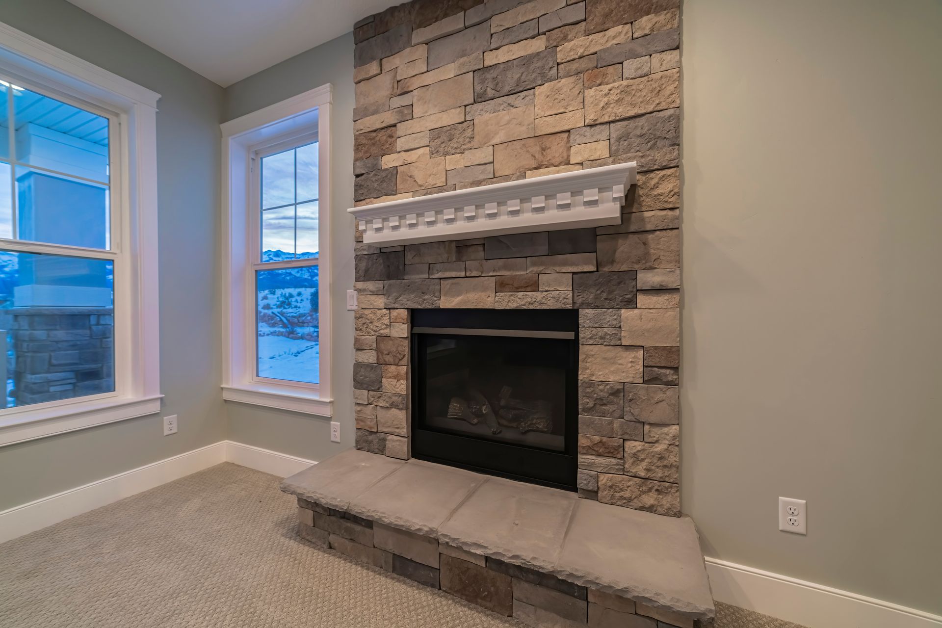 Fireplace with stone surround, mantle, and cushioned bench. Windows on left. Beige carpet and wall. Fireplace with stone surround, mantle, and cushioned bench. Windows on left. Beige carpet and wall.