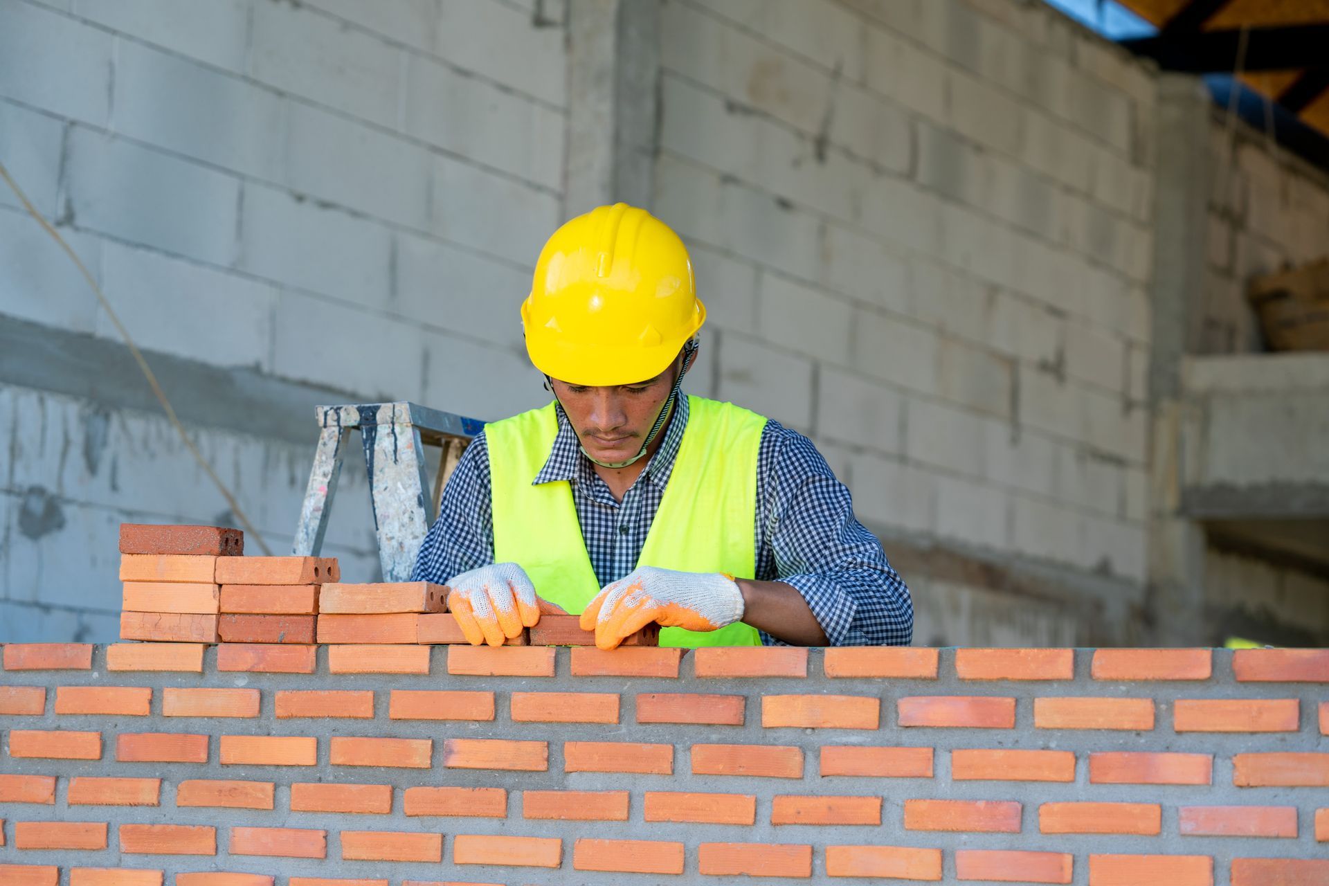 Construction worker laying bricks; yellow hard hat, reflective vest, outdoor setting.