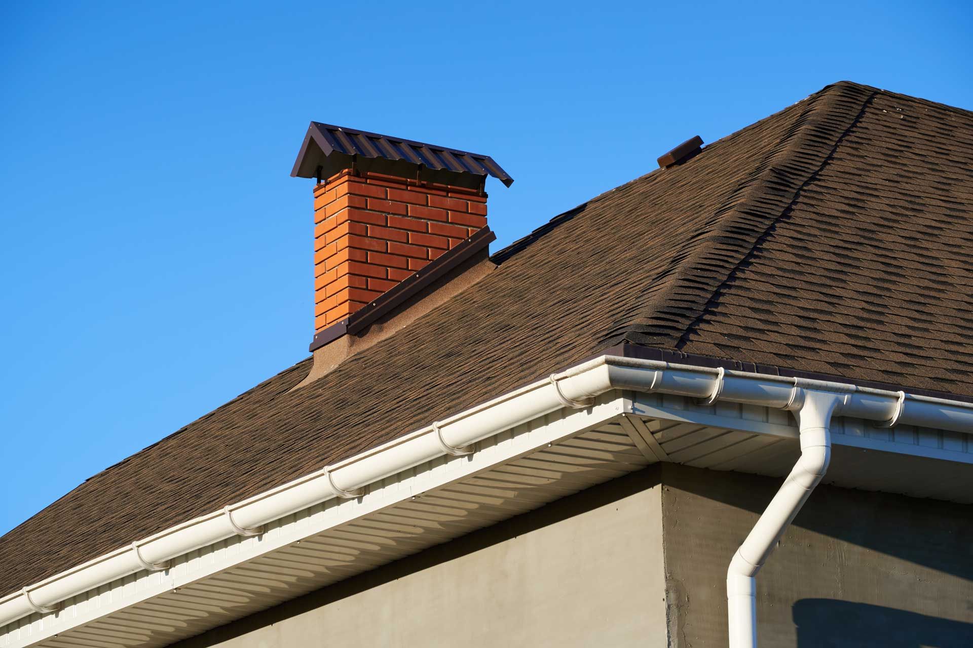 Brown shingled roof with white gutters and a red brick chimney against a blue sky.