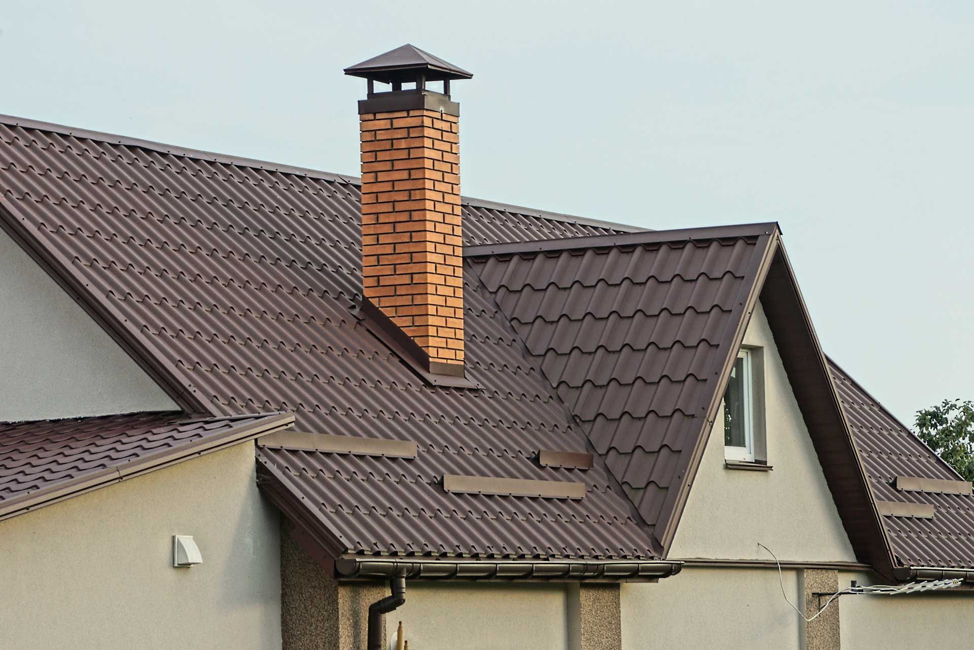 Brown metal roof with brick chimney on a light-colored building under a pale sky.