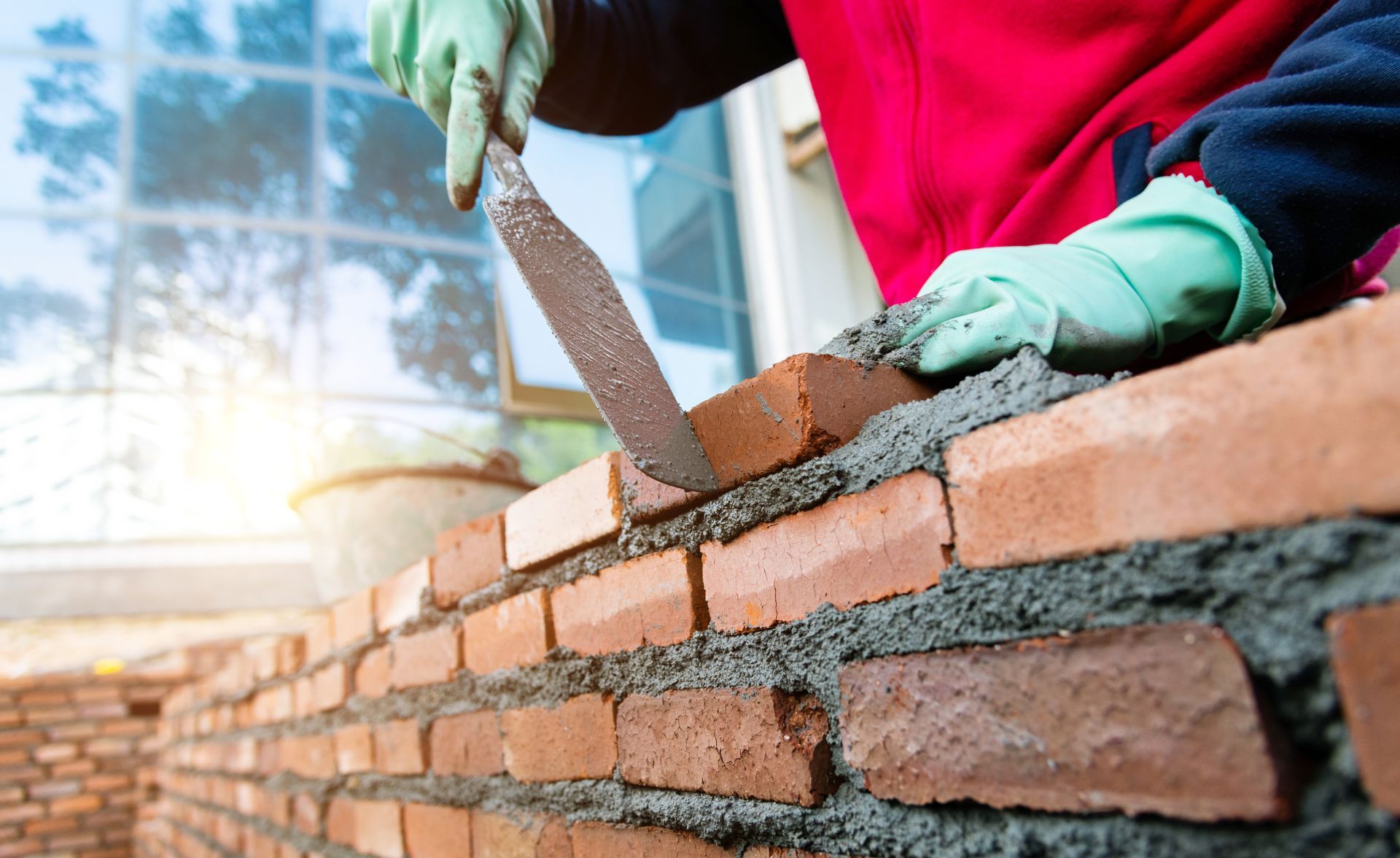 Person in green gloves uses a trowel to apply mortar on brick, building a wall.